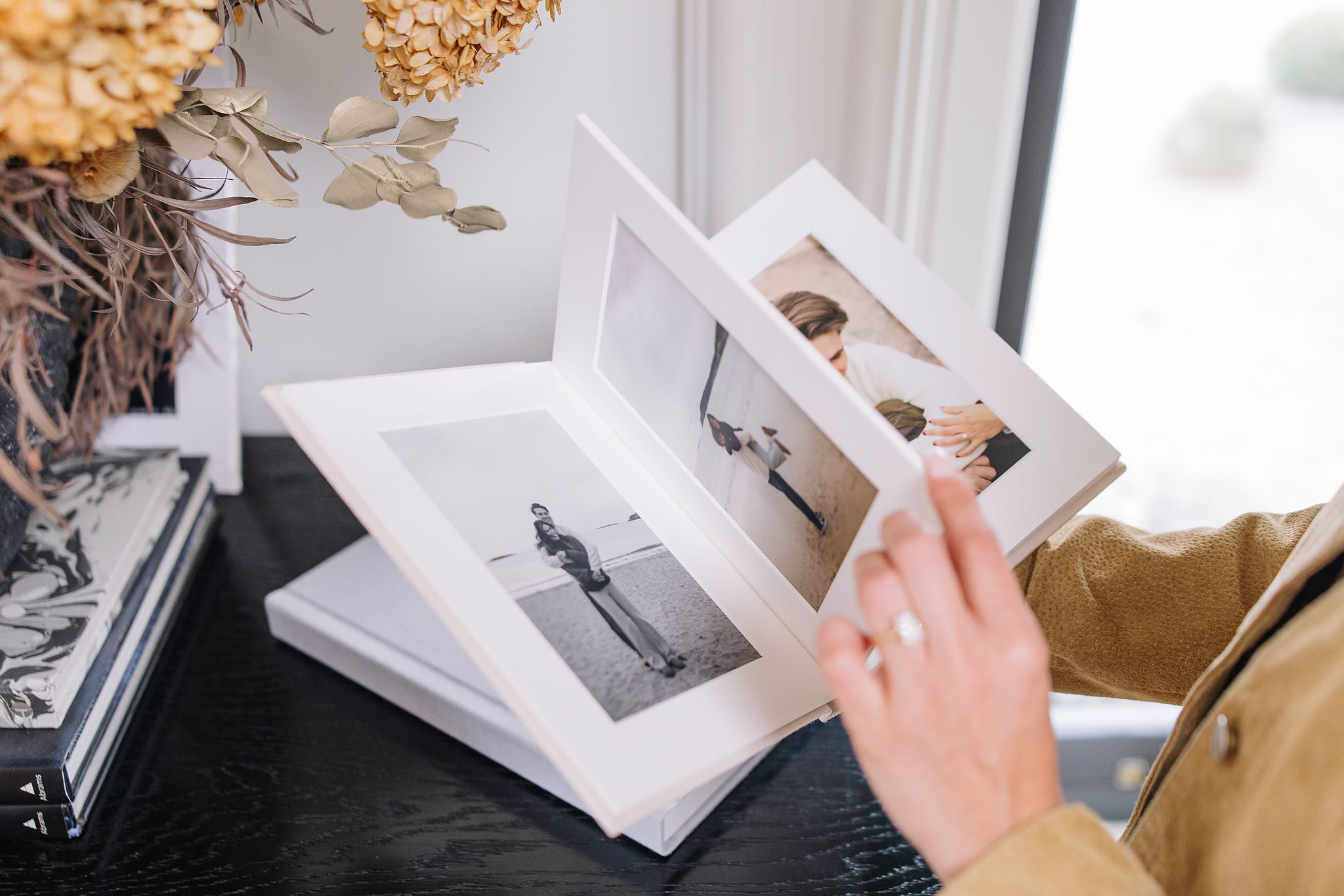 A hand lifts a matted page in an open white linen folio album, an engagement ring on her finger and dried hydrangeas above